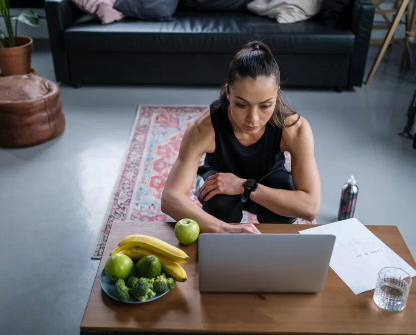 A woman in athletic wear sits at a low table in her living room, looking at a laptop. On the table are fruits, vegetables, a glass of water, a water bottle, and diabetessisters notes to support her healthy lifestyle.