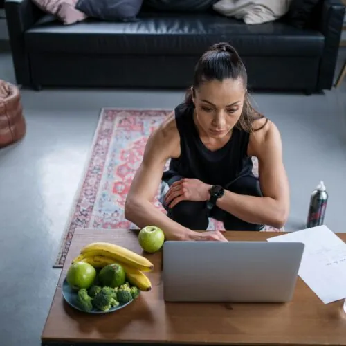 A woman in workout clothes sits on the floor in front of a laptop on a coffee table with fruit, vegetables, a water bottle, and papers, embracing a healthy lifestyle inspired by diabetessisters, in a living room with a sofa and plants.