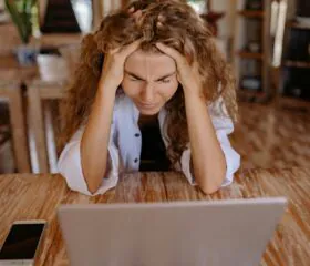 A woman with curly hair sits at a table, holding her head in her hands and looking stressed while staring at a laptop. A smartphone lies on the table beside her in a warmly lit room.