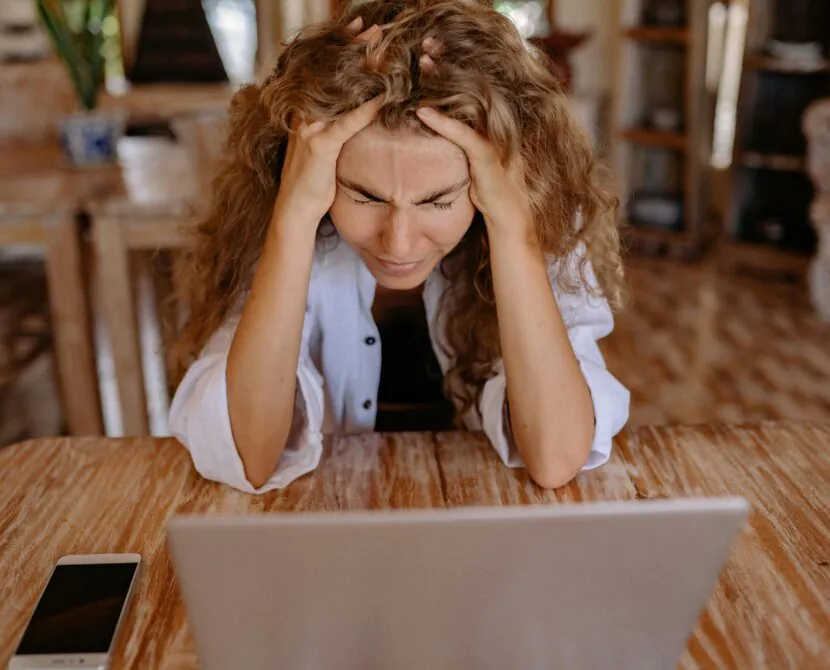 A woman with curly hair sits at a table, holding her head in her hands and looking stressed while staring at a laptop. A smartphone lies on the table beside her in a warmly lit room.