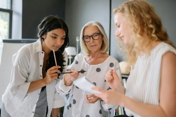 Three women, part of DiabetesSisters, stand together in an office discussing notes. One holds a notepad while another points, and the third listens attentively with a pen. Shelves and a flip chart are visible in the background.