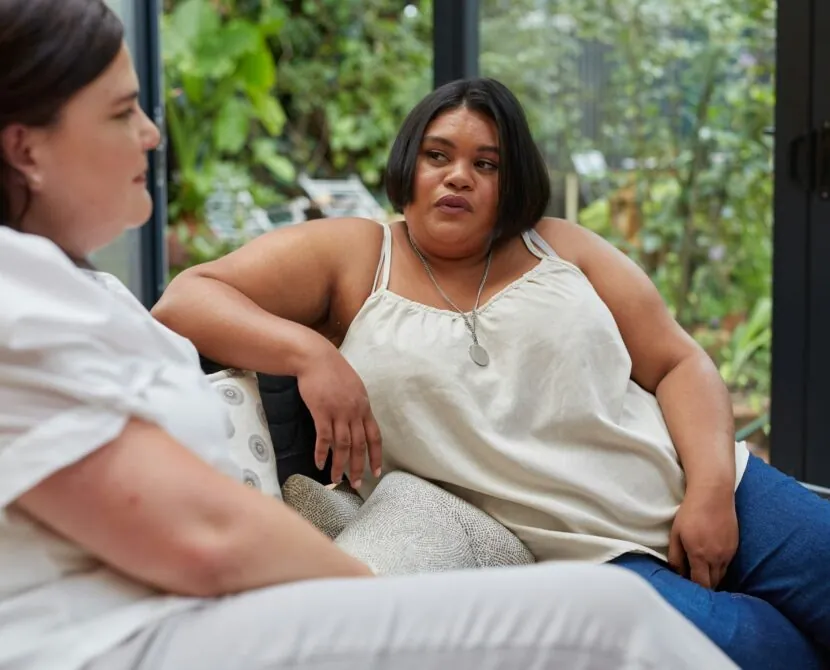 Two women sit on a couch indoors, having a serious conversation about weight loss medications and diabetes. One leans back, listening attentively as the other speaks. Green plants are visible outside through large windows in the background.