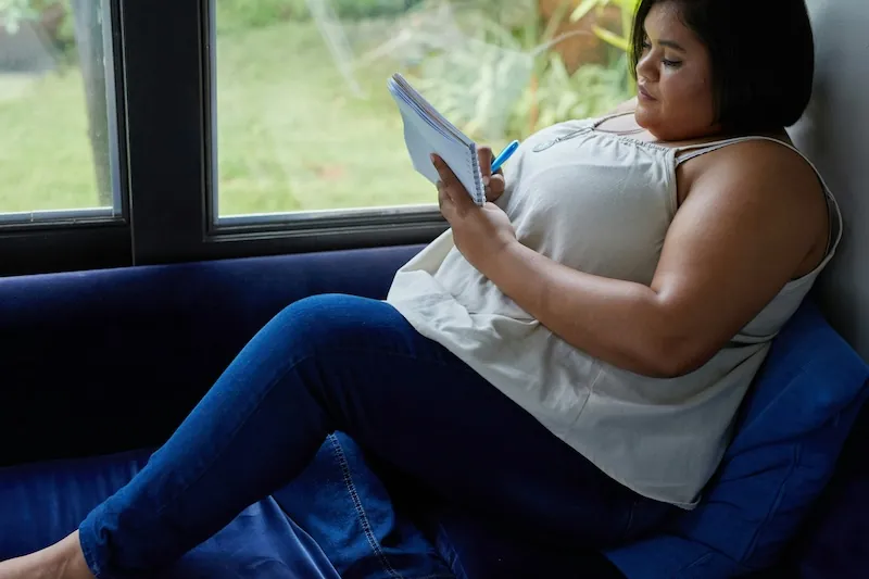 Woman sitting on a couch writing in a notebook, reflecting on her health journey with diabetes and weight care options.