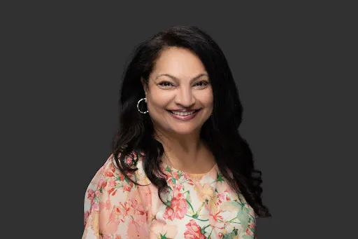 A woman with long dark hair, wearing hoop earrings and a floral-patterned blouse, smiles at the camera against a plain dark gray background.